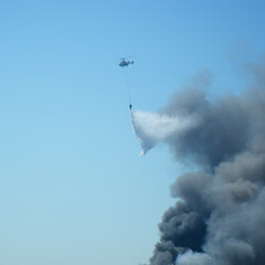 Aerial firefighting with helicopter. Black smoke from the fire over the city in the blue sky.