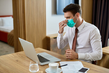 Male entrepreneur adjusting face mask while using laptop in hotel room.