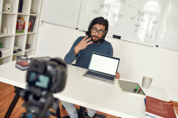 Fototapeta premium Young male teacher sitting at his workplace and explaining new theme while recording video blog on professional digital equipment. Coding class online