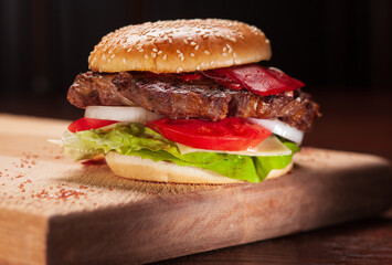Tasty burger on wooden cutting board. Hamburger with pieces of fried meat, red bell pepper, onions and lettuce leaves. Selective focus