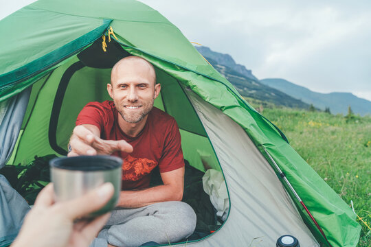 Smiling bald Man in the green tent taking the thermos teacup for morning tea-drinking with mountain background. Active people enjoying traveling with tents concept image.