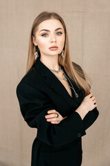 A beautiful girl of European appearance with brown straight loose hair, with neutral makeup, in a black jacket, arms crossed on her chest. Beige background.Vertical orientation.