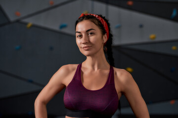 Close up of active young woman in sportswear looking at camera, standing against artificial training climbing wall. Concept of sport life and rock climbing