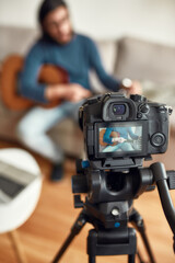Guitar lessons. Young man sitting on sofa at home and teaching how to play guitar. Recording webinar at home. Learning music online. Focus on a camera