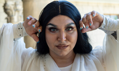 Drag queen posing close to old building in white dress. A wig with Black long hair and make up. High quality photo