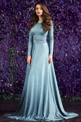 Obraz na płótnie Canvas Young beautiful elegant woman in blue long evening dress posing against wall of purple flowers. Spring fashion style portrait of gorgerous girl