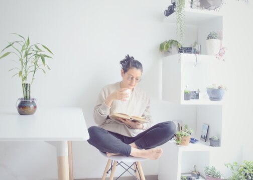 Hermosa mujer relaj&aacute;ndose en su casa bebiendo caf&eacute; leyendo un libro en la ma&ntilde;ana. Mujer empieza el d&iacute;a con un caf&eacute; y un libro en la sala de su casa.