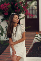 Young beautiful stylish trendy woman in white dress sitting and relaxing in garden near small white house