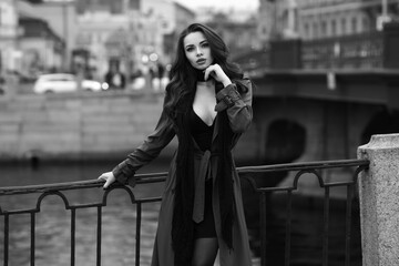 Pretty woman wearing stylish black dress with decollete, scarf and coat posing at embankment in city centre