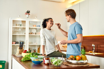 Obraz na płótnie Canvas Happy couple, vegetarians preparing healthy meal, sandwhich, salad in the kitchen together. Guy feeding his girlfriend dried fruits. Vegetarianism, healthy food, diet concept