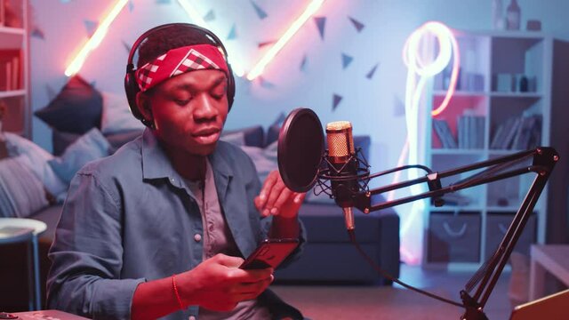 Young cool African American man in headphones holding smartphone and reading rap in condenser microphone while recording hip-hop music in home studio with neon light