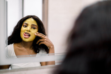 Portrait of young smiling African brunette woman in white t-shirt, standing in bathroom at home, looking in the mirror and applying golden antiwrinkle mask on her face. Back reflection