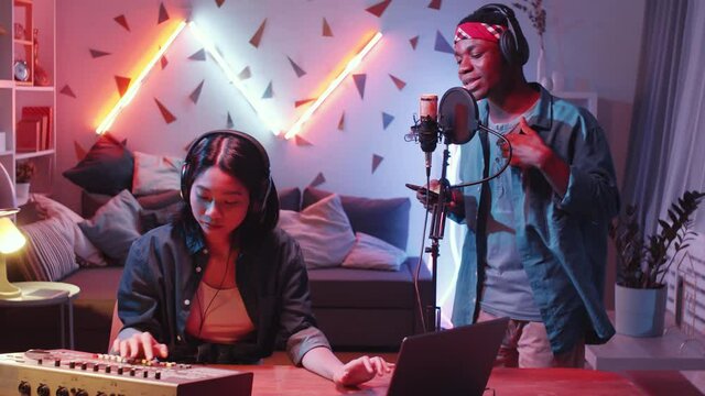 Multiethnic music duo recording hip-hop at home studio: cool African American man in headphones holding smartphone and reading rap in microphone while Asian female dj using mixing console at desk
