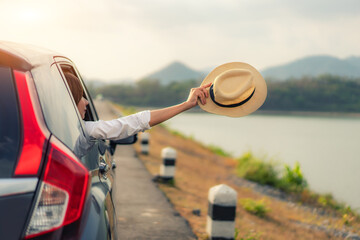 Woman at spree time relaxing and enjoying road trip. Young Asian happines female inside compact black car with raise her hat out to the car window with mountain and lake view at sunny day in Thailand.