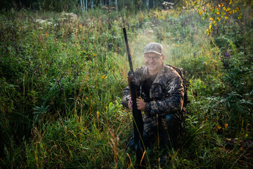 The smiling hunter or military person with gun or rifle in summer forest during the bird hunt.