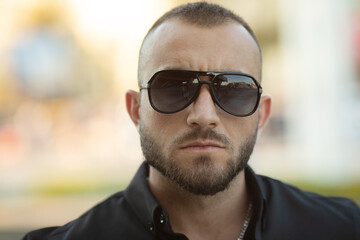 Confident handsome man in sunglasses and shirt over city background. Men's beauty, fashion. Outdoor portrait.