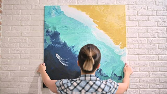 the girl hangs an oil painting on the wall and admires her. View from the back