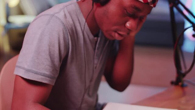 Close up shot of young African American hip-hop producer in headphones sitting at desk in home recording studio, moving his head to music and writing lyrics in notepad while working on new song