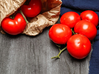 ripe tomatoes on a twig lie in a paper bag on a wooden wooden table and next to a blue kitchen napkin, closeup side view.