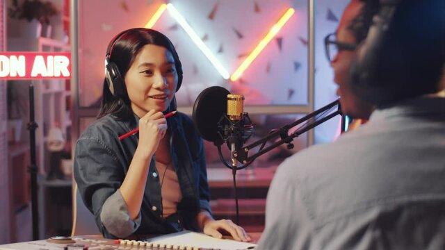 Over the shoulder shot of young Asian female host in headphones having conversation with Afro-American male guest while broadcasting radio interview in studio
