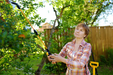 Middle age caucasian woman sprays trees against pests, vermin or diseases plants. Seasonal working in garden.