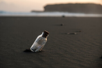 Glass bottle on a black sand beach at sunset. A romantic message in a bottle. Glass jar with shells on the beach.