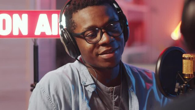 Over the shoulder shot of young African American male radio host in headphones interviewing guest while broadcasting in studio with lighted on-air sign