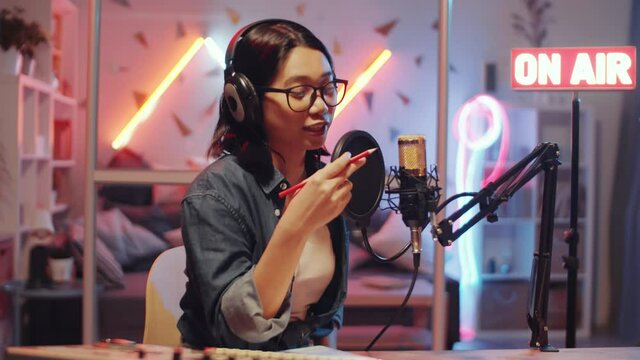 Young Asian woman putting headphones on and talking into microphone while recording podcast in home radio studio with colorful neon light and illuminated on-air sign