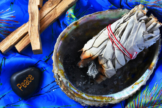 And image of white sage bundles in an abalone shell on a bright blue silk background. 