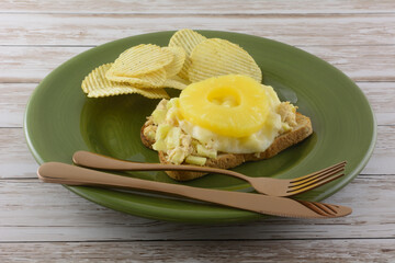 Open face tuna salad sandwich with melted provolone cheese slice and pineapple fruit ring with potato chips on green plate with knife and fork