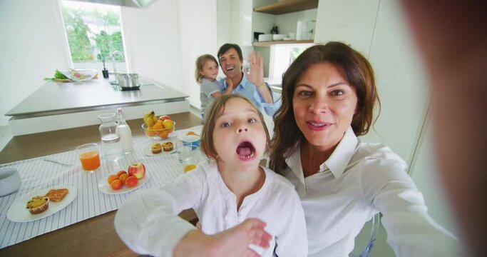 A happy smiling family is making a selfie or video call with to friends or relatives while having a breakfast together in a kitchen at home in the morning.
