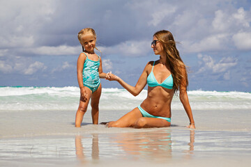 Beautiful sexy young caucasian woman in blue bikini and little girl on the sandy beach. Summer family vacations, travel and tourism concept after end of coronavirus covid-19 lockdown. Copy space