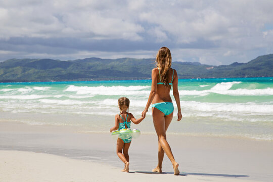 Beautiful sexy young caucasian woman in blue bikini and little girl on the sandy beach. Summer family vacations, travel and tourism concept after end of coronavirus covid-19 lockdown. Copy space