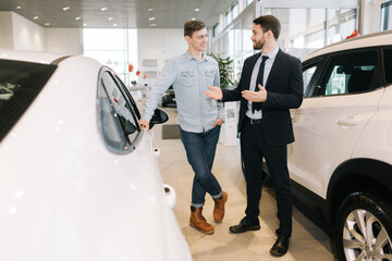 Professional car salesman wearing business suit showing new car interested buyer in auto dealership. Concept of choosing and buying new car at showroom