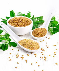 Fenugreek in two spoons and bowl with leaves on wooden board
