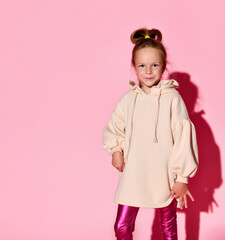 Cute little girl with updo posing with confident look in pale-pink sweatshirt and glossy leggings. Three quarter length isolated on pink, copy space