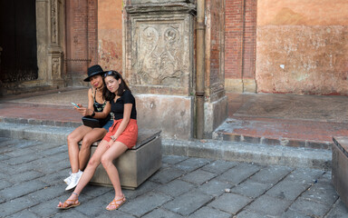 Fototapeta na wymiar Mother and daughter with red shorts sitting on bench on road looking at photographer while holding cell phone