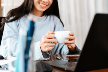 woman working on laptop and hold cup of coffee while sitting at working place.