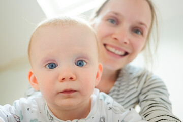 Serious adorable blue eyed baby looking at camera, blurred mom smiling in background. Closeup shot, front view. Child care or childhood concept