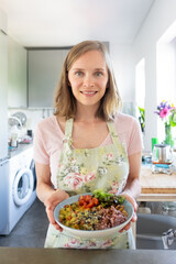 Food blogger with veggy bowl. Happy woman holding homemade vegetable dish, standing in kitchen, looking at camera and smiling. Vertical shot. Vegetarian diet concept