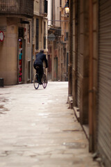 A man is cycling in a narrow street among houses and shops on a cobblestone ground. Image was taken in old town in Barcelona, Spain A boutique as well as shutters of closed shops are in the background