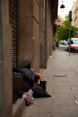 An elderly man is sleeping on a doorway by the metal shutters of a shop during day time. He wears boots and a black coat. Sidewalk, cars, trees and buildings are seen in the background.