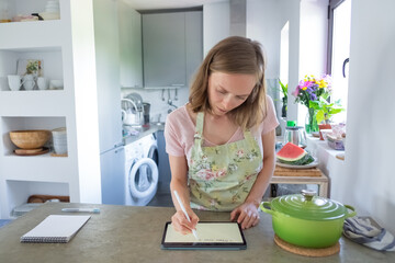 Focused young woman planning weekly menu, writing on pad screen while cooking in her kitchen, using tablet near big saucepan on counter. Front view. Cooking at home and household concept