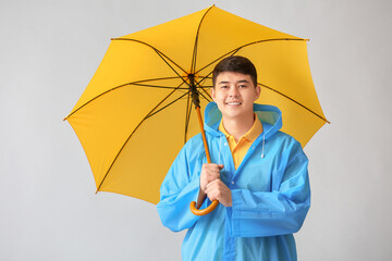 Young Asian man in raincoat and with umbrella on grey background