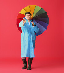 Young Asian man in raincoat and with umbrella showing OK on color background