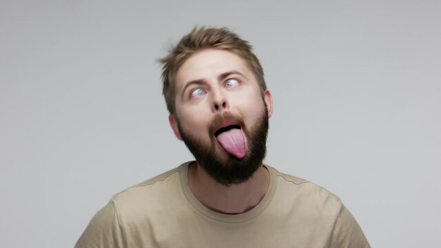 Brainless dumb cheerful bearded guy showing tongue out, looking cross eyed and aping as fool, behaving childish disobedient, silly humorous expression. indoor studio shot isolated on gray background