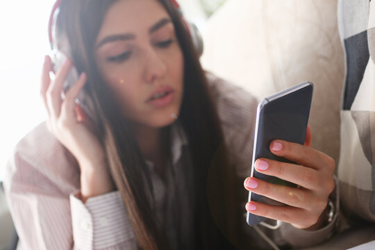 Young beautiful woman headphones holds smartphone in hand using at home costs about sofa. Verifies internal information its financial advertising freedom makes money level transfer contractor concept