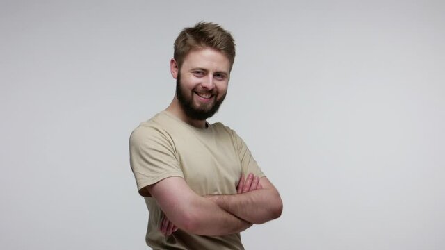 Cheerful handsome bearded guy turning crossed hands and looking at camera with toothy smile, feeling confident self-assertive, optimistic outlook. indoor studio shot isolated on gray background