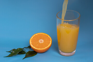 Fresh orange juice is poured into a glass, fruit cut in half and sliced in green leaves isolated on a blue background