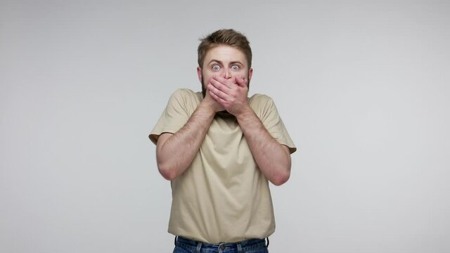 Scared bearded guy looking at camera with big eyes shocked expression, closing mouth zipping lips to keep terrible truth secret, feeling intimidated. indoor studio shot isolated on gray background